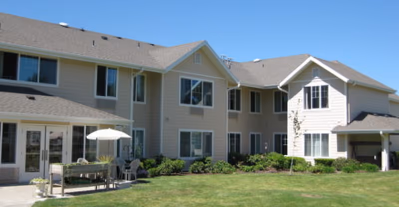 Front exterior of a two-story beige senior living building with a lawn and a small patio table and umbrella.