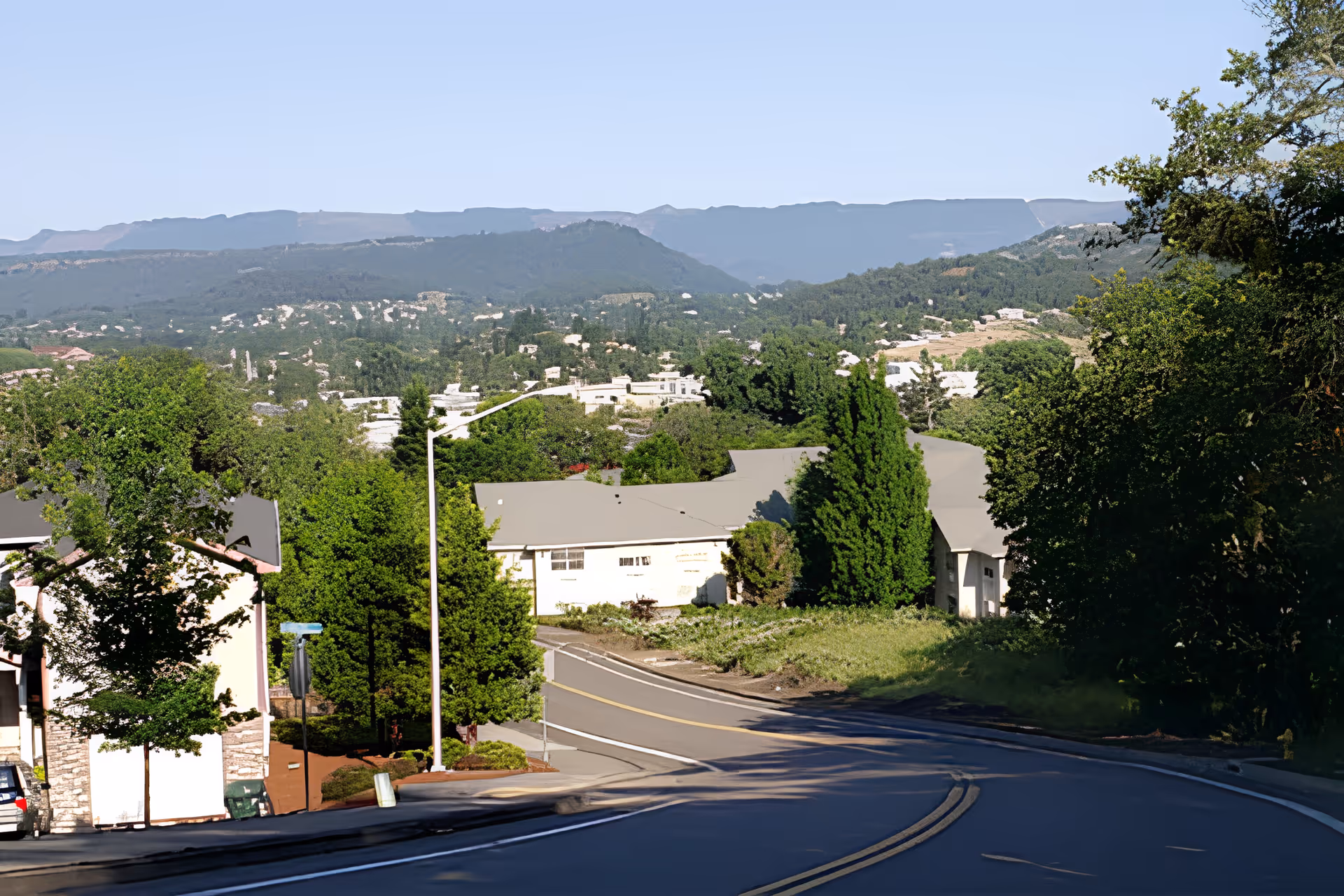 A winding downhill street lined with trees and houses, with a valley of buildings and distant mountains under a clear sky.
