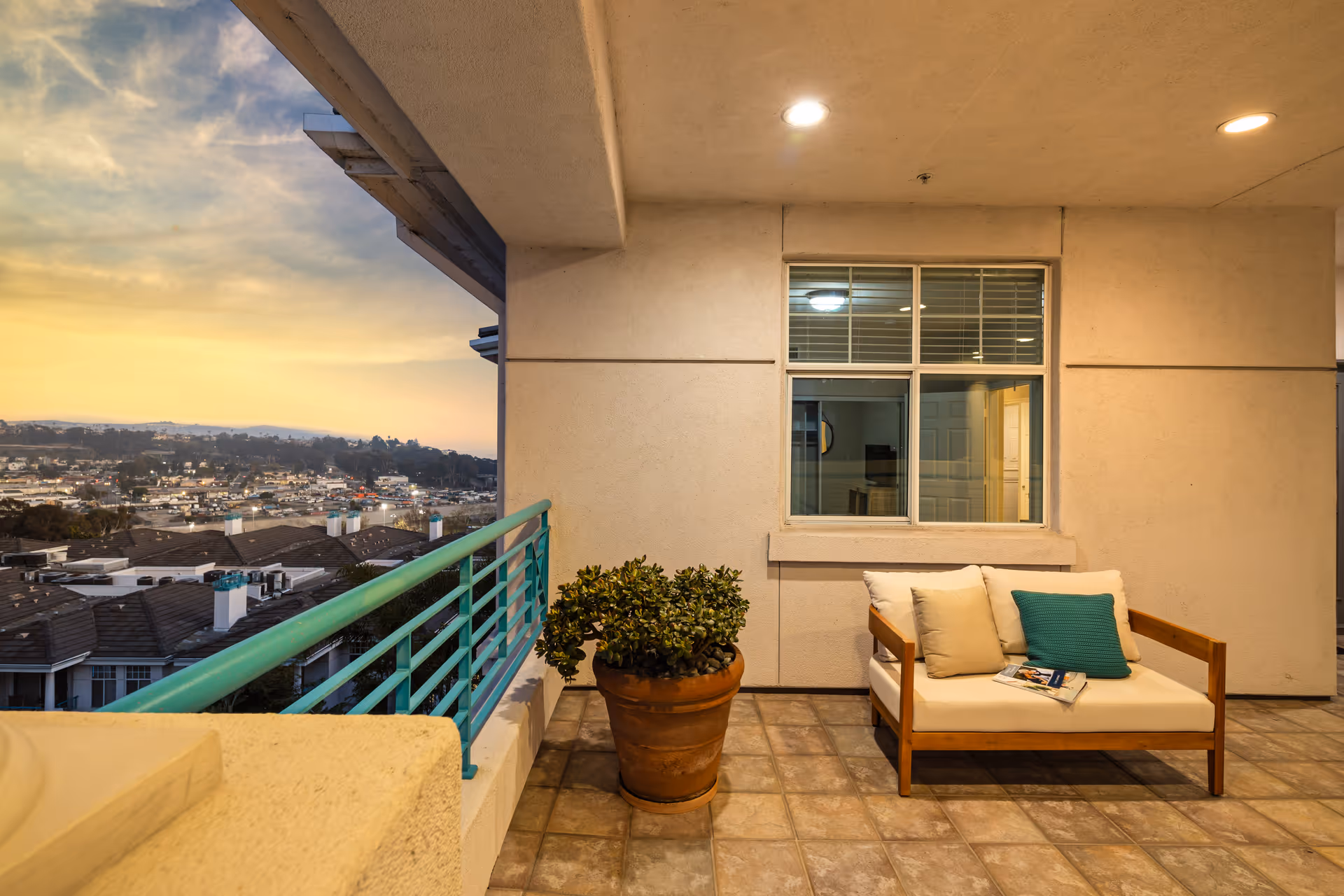 Outdoor balcony area at sunset with a cushioned wooden loveseat featuring beige and teal pillows, a large potted plant, tiled floor, and a view of rooftops and distant hills under a partly cloudy sky.