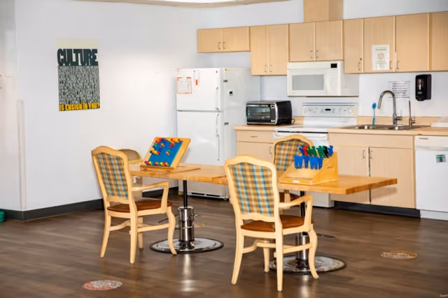 A communal kitchenette and dining area with wooden tables, plaid-upholstered chairs, a refrigerator, microwave, sink, and activity materials on the tables.