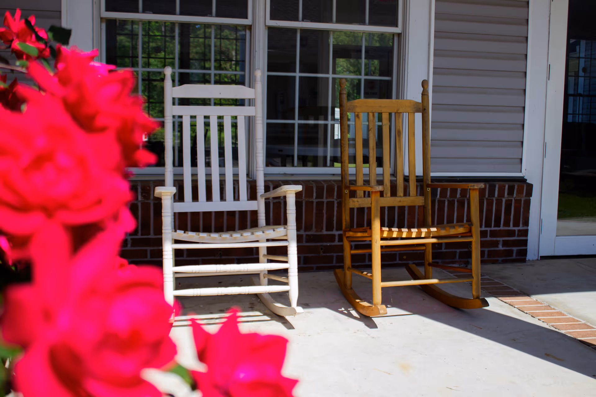 Two wooden rocking chairs sit on a sunlit porch in front of a building with bright red flowers in the foreground.