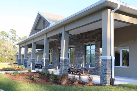 Exterior view of a senior living facility with a covered porch supported by stone pillars. Several rocking chairs are placed along the porch, and there is a landscaped garden with small plants and mulch in front. The building has large windows and a light-colored roof.