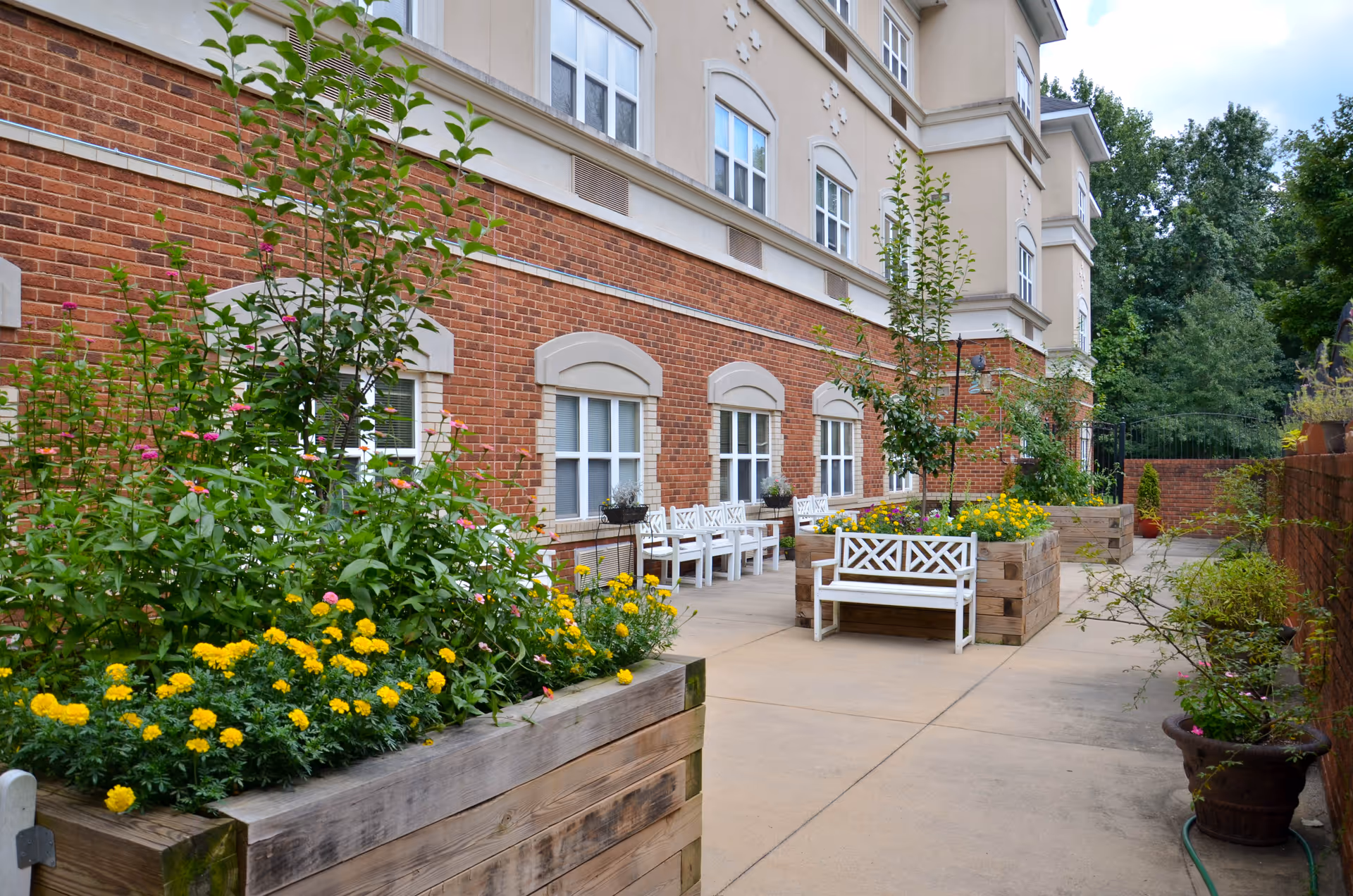 Outdoor courtyard with raised wooden planters filled with flowers and white benches along a brick-faced building.
