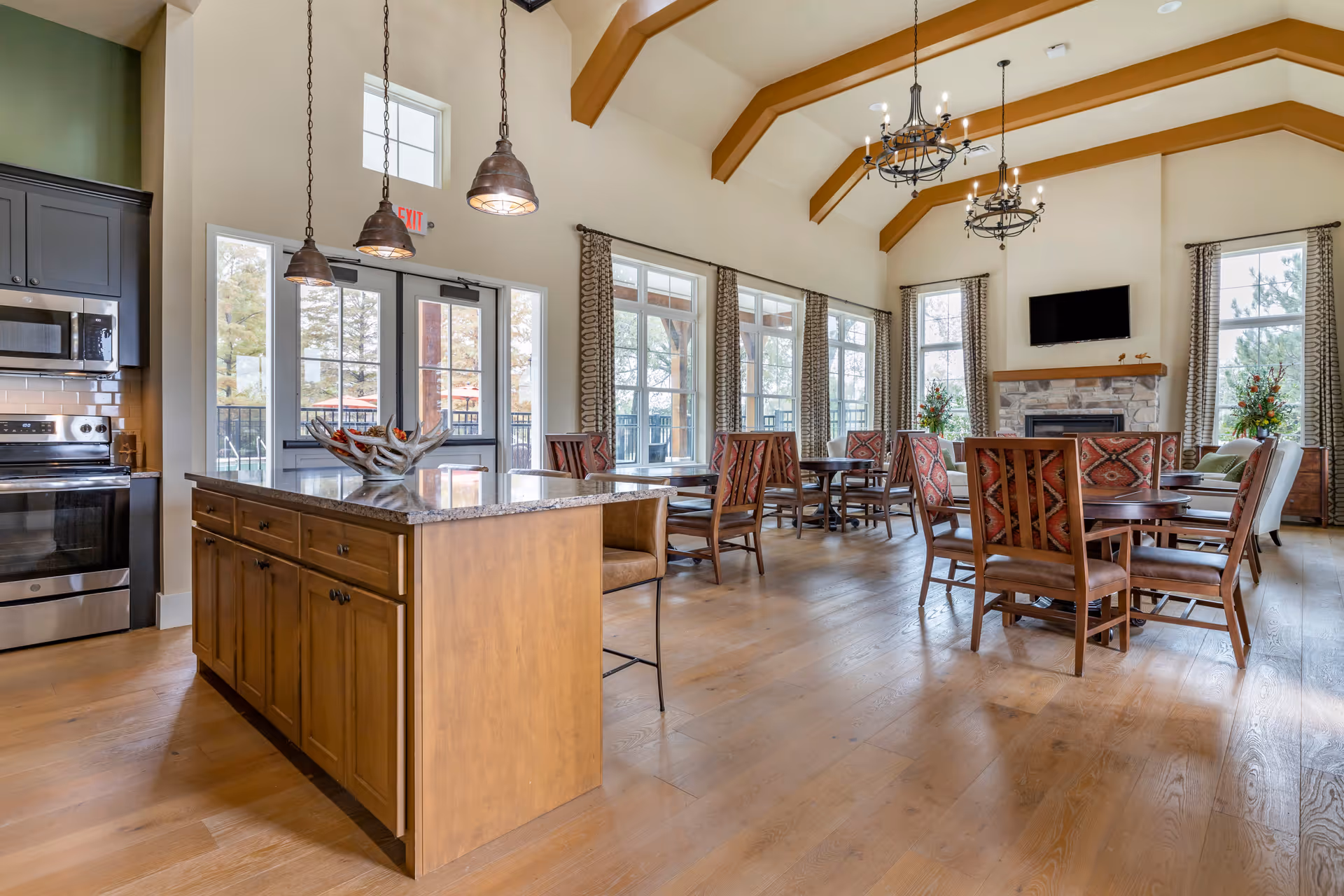 A spacious senior living common area featuring a kitchen island with pendant lights, wooden dining tables with patterned chairs, large windows with curtains, a stone fireplace with a mounted TV above, and exposed wooden ceiling beams.