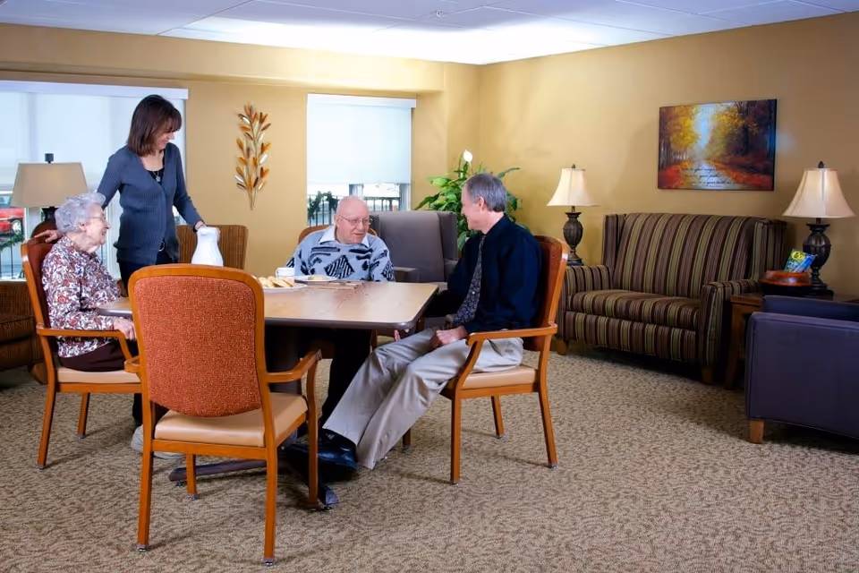 A staff member and three elderly residents sit and talk around a table in a comfortable senior living lounge with chairs, a striped sofa, lamps, and wall art.