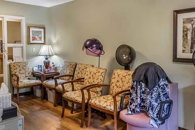 A cozy waiting area with four patterned armchairs lined up against a light green wall. Two hair drying stations with hooded dryers are positioned near the chairs. A small wooden side table with a lamp and framed pictures is placed between two of the chairs. The floor is wooden, and a framed picture hangs on the wall.