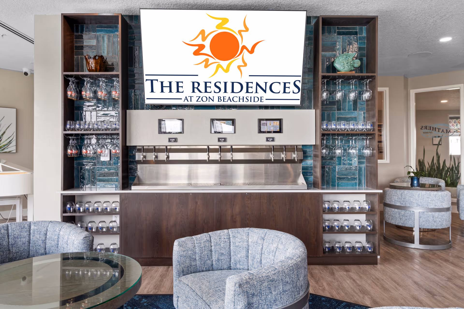 Interior view of a lounge area at The Residences at Zon Beachside featuring a beverage tap system with multiple taps, shelves stocked with glasses, and comfortable blue upholstered chairs around a glass table.