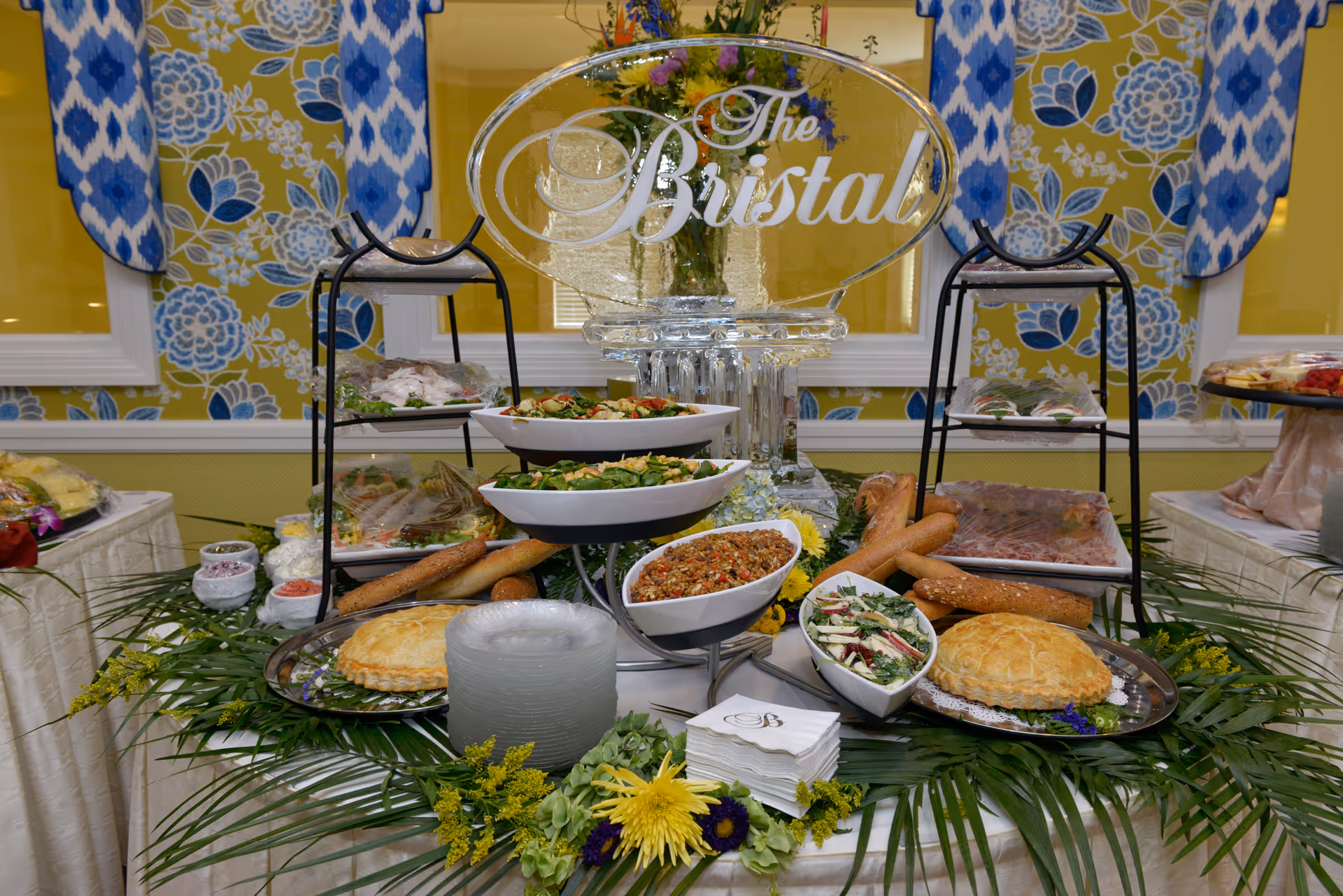Buffet table with salads, breads, pies and an ice sculpture reading "The Bristal" in a decorated dining room.