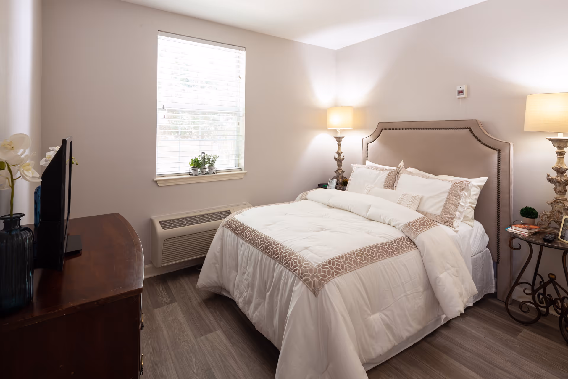 A cozy bedroom with a neatly made bed featuring white and beige bedding. The room has a window with blinds, a wooden dresser with a TV on top, two bedside tables with ornate lamps, and a small plant on one table. The walls are painted light beige and the floor has wood-like flooring.