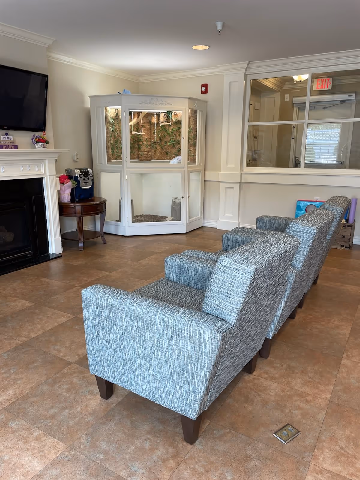 A cozy living room area with three gray upholstered armchairs arranged in a row facing a fireplace with a mounted TV above it. To the left of the fireplace is a small round wooden table with a coffee machine and some decorative items. In the corner, there is a white enclosed terrarium or display case with plants inside. The room has beige tiled flooring and cream-colored walls with white trim. A window and an exit door are visible in the background.