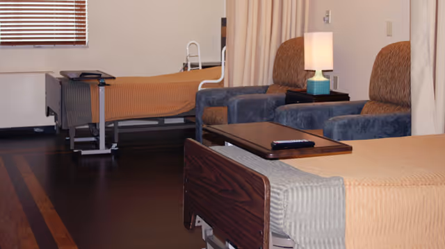 Interior view of a senior living facility room with two hospital-style beds, each covered with a beige blanket. Between the beds are two upholstered armchairs and a small table with a lamp. A window with blinds is visible on the left side.