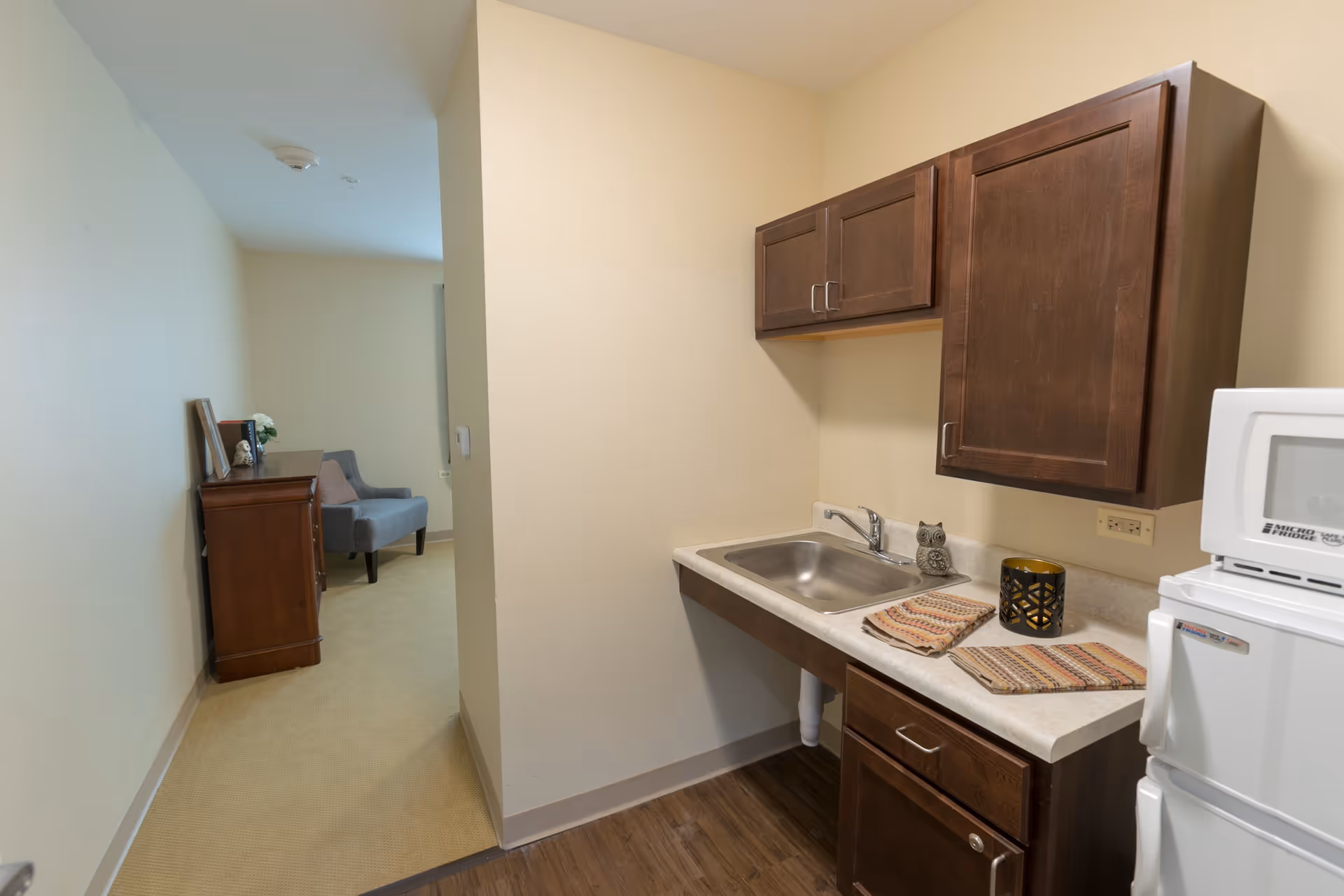 Small kitchenette area with a stainless steel sink, dark wood cabinets, a microwave on top of a mini refrigerator, and two patterned cloths on the countertop. In the background, there is a hallway leading to a room with a wooden dresser and a gray upholstered chair.