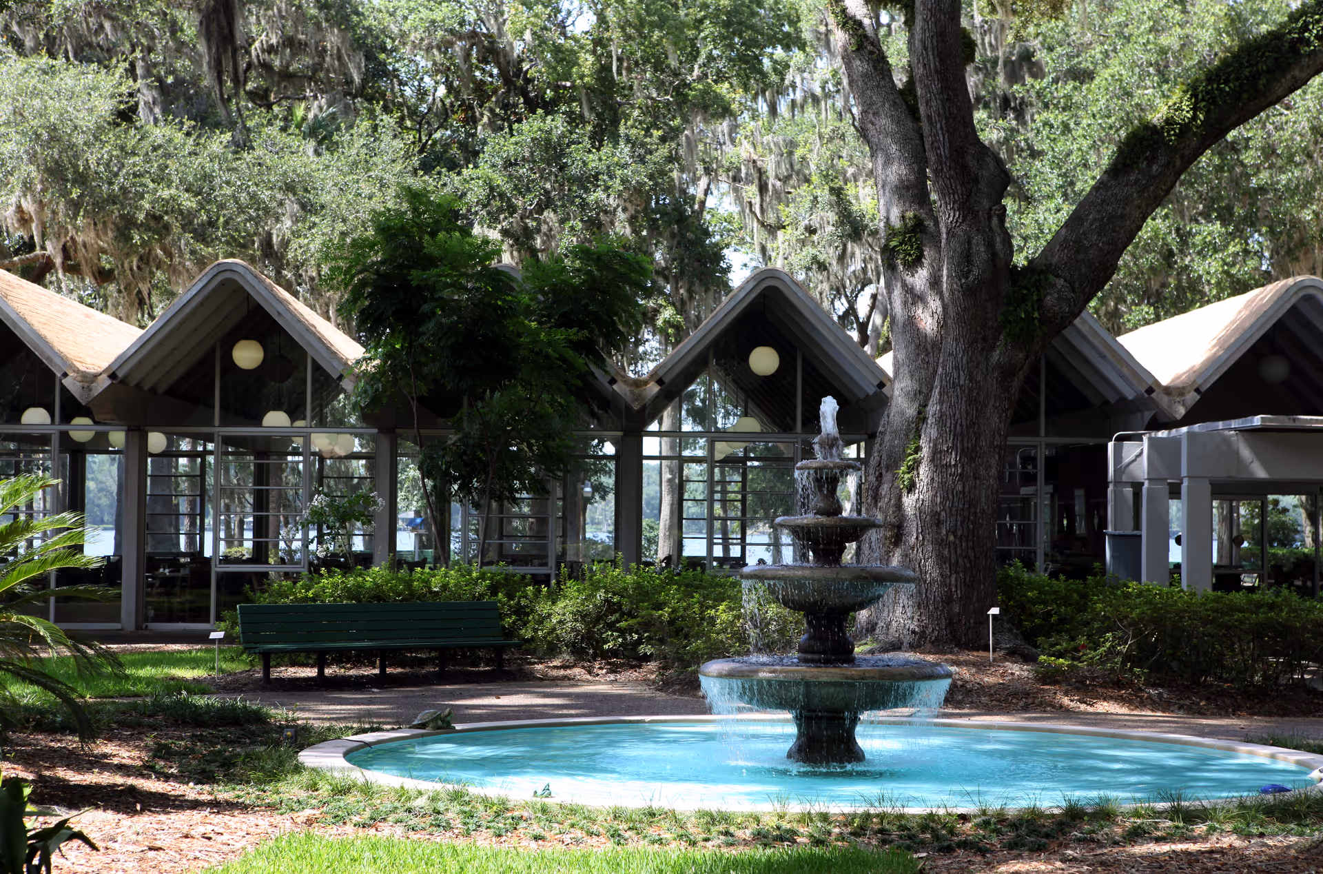 Outdoor scene at Westminster Woods on Julington Creek featuring a multi-tiered water fountain in the foreground surrounded by greenery and a large tree. Behind the fountain is a building with large glass windows and a distinctive zigzag roof design, with trees and a body of water visible in the background.
