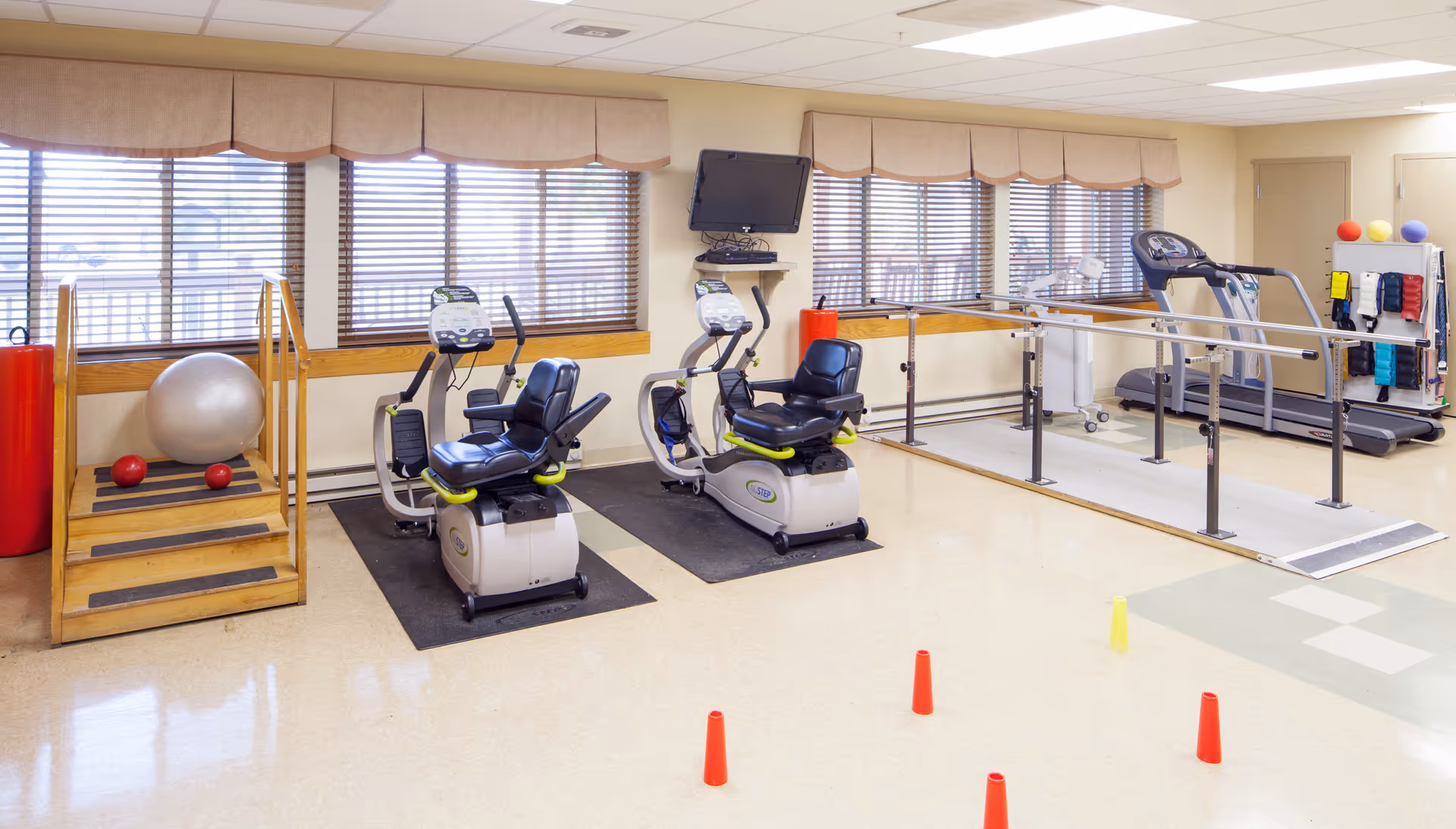 Bright interior therapy/exercise room with recumbent bikes, a treadmill, parallel bars, stairs and small cones on the floor.