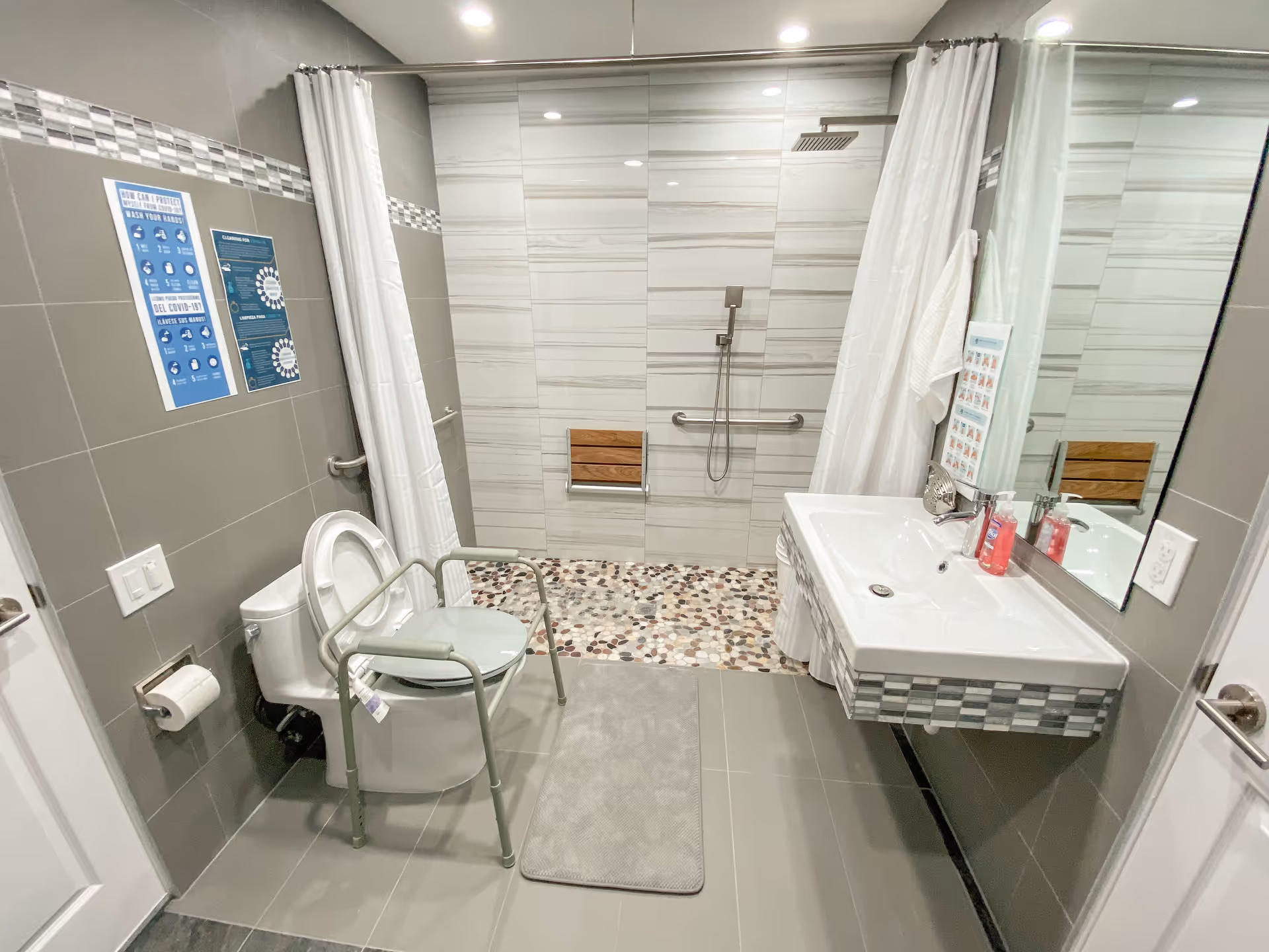 Accessible senior bathroom with a walk-in tiled shower, toilet with a transfer bench, wall-mounted sink, and grab bars.