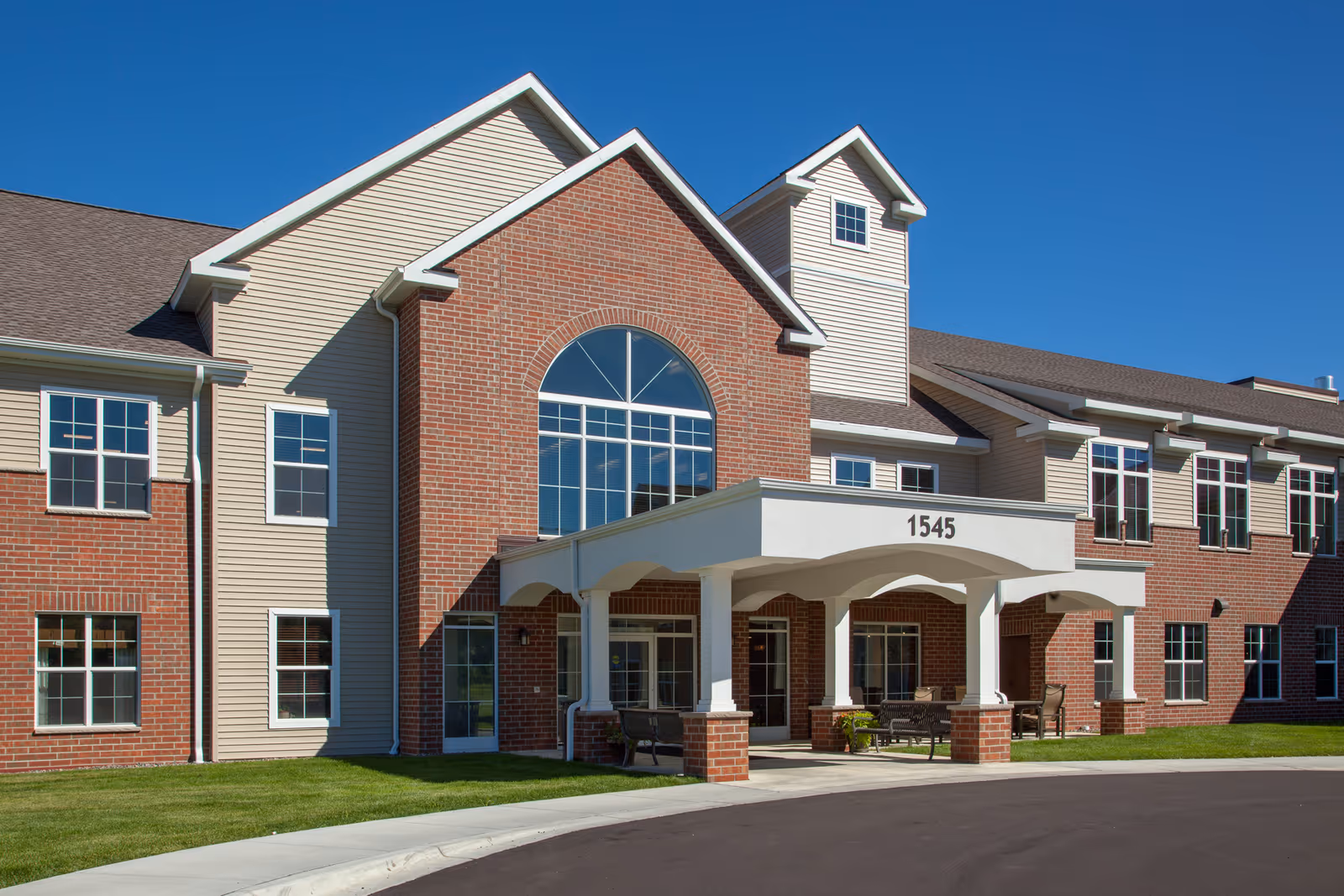 Exterior view of a senior living facility named GracePointe Crossing, featuring a two-story building with a combination of red brick and beige siding, multiple windows, a covered entrance with the number 1545 displayed, and a clear blue sky above.