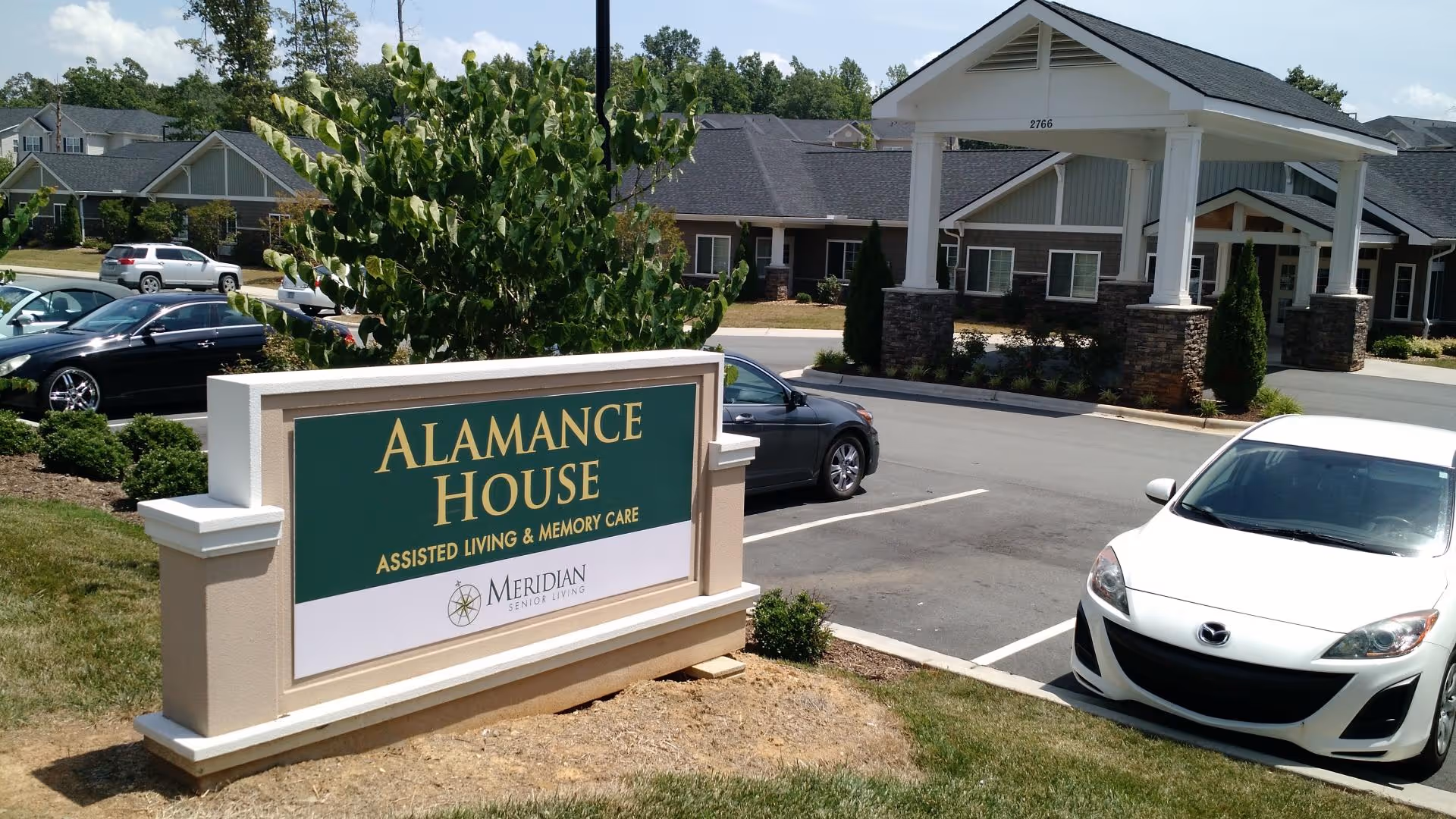Entrance sign that reads 'Alamance House Assisted Living & Memory Care' in front of the facility building and parking lot.