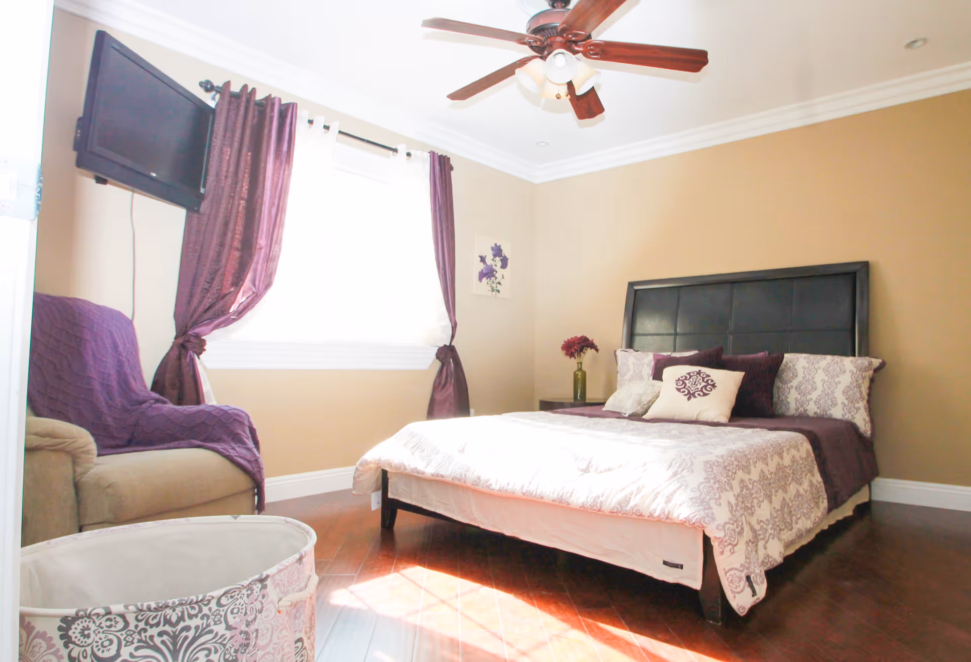 A bright bedroom with a large bed featuring a black headboard, white and purple bedding, and decorative pillows. There is a beige armchair with a purple throw blanket near a window with purple curtains. A ceiling fan with wooden blades is mounted on the white ceiling. A small side table with a vase of flowers is next to the bed, and a wall-mounted TV is visible near the window.