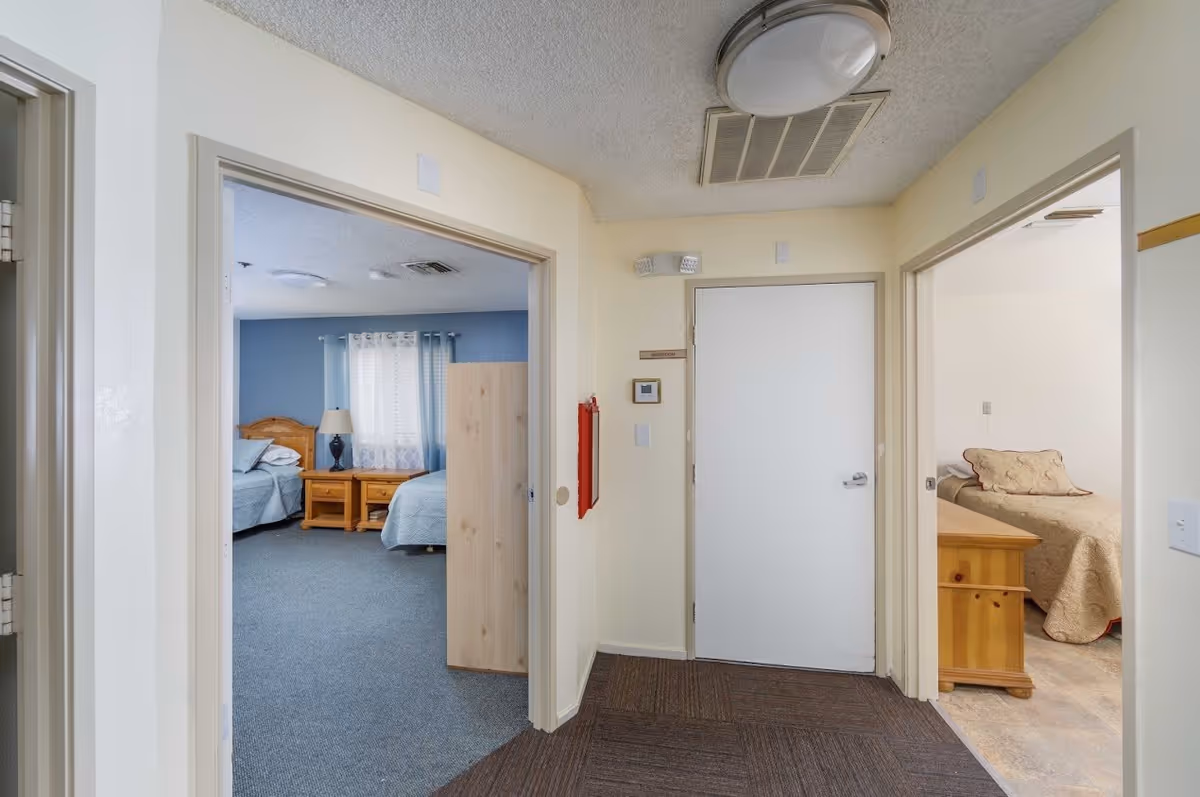 View of a hallway in a senior living facility with two open doorways leading to bedrooms. The left bedroom has two beds with blue bedding, a wooden nightstand, and a lamp. The right bedroom has a single bed with beige bedding and a wooden chest at the foot of the bed. The hallway has a white door labeled 'Restroom' and a red fire alarm box on the wall.