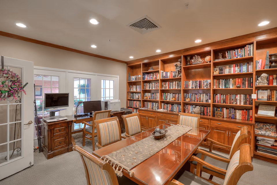 A cozy interior library/meeting room with built-in wooden bookshelves, a polished wooden table surrounded by striped chairs, and a desk with a computer near French doors.