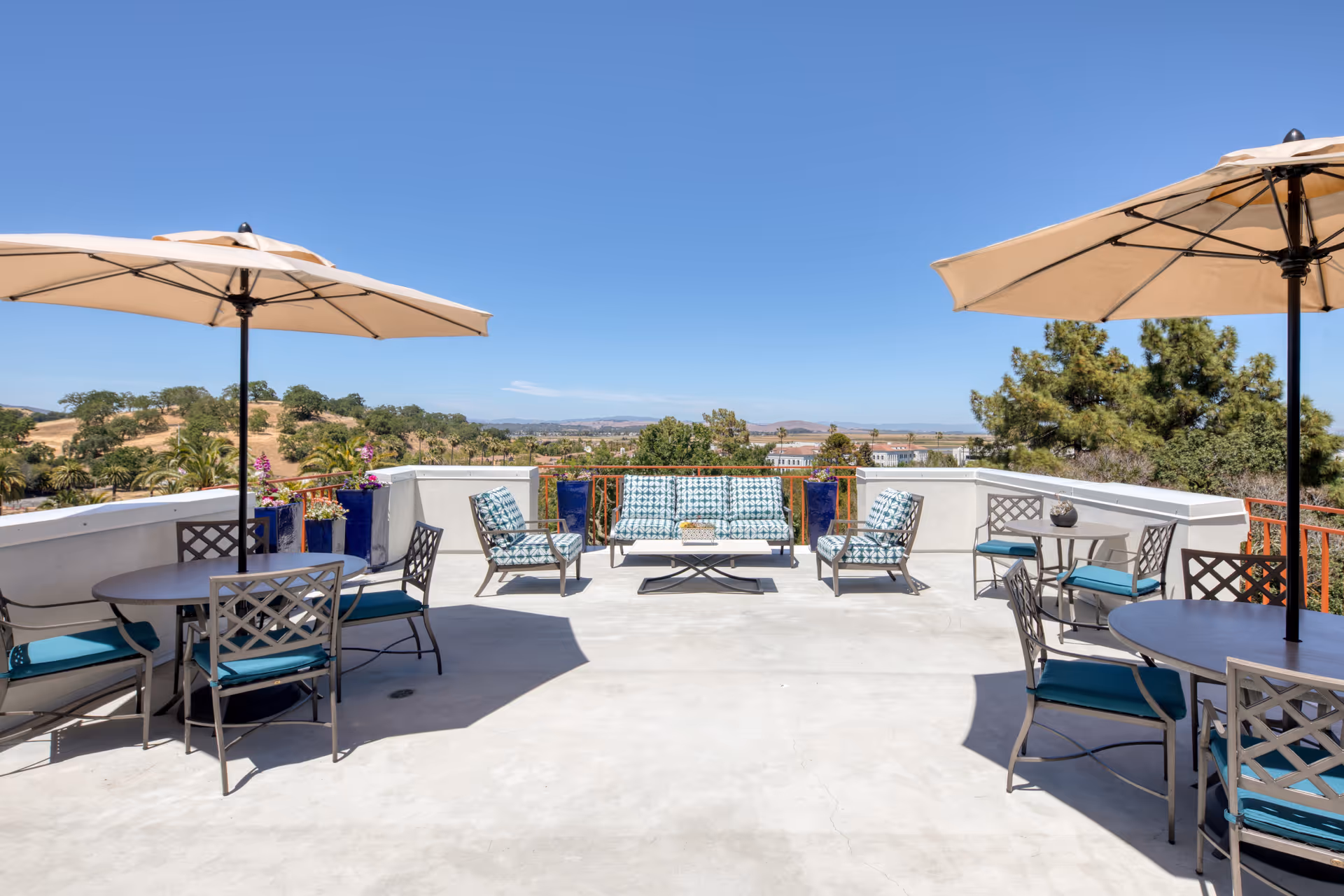 Outdoor patio area with multiple seating arrangements including cushioned chairs and sofas, round tables with umbrellas providing shade, overlooking a scenic view of trees, hills, and distant buildings under a clear blue sky.
