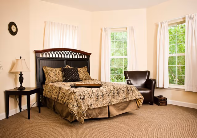 A cozy bedroom with a large bed featuring a patterned bedspread and multiple pillows. The room has beige walls, carpeted floor, two large windows with white curtains, a dark wooden headboard, a black leather armchair, a small side table with a lamp, and a wall clock.