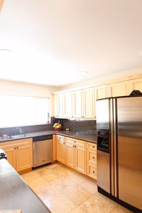 Bright kitchen with light wood cabinets, stainless steel refrigerator and dishwasher, black countertops, and a window above the sink letting in natural light.