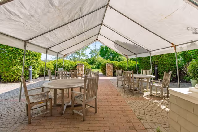 Outdoor seating area with round wooden tables and chairs under a large white canopy tent, surrounded by green hedges and trees.