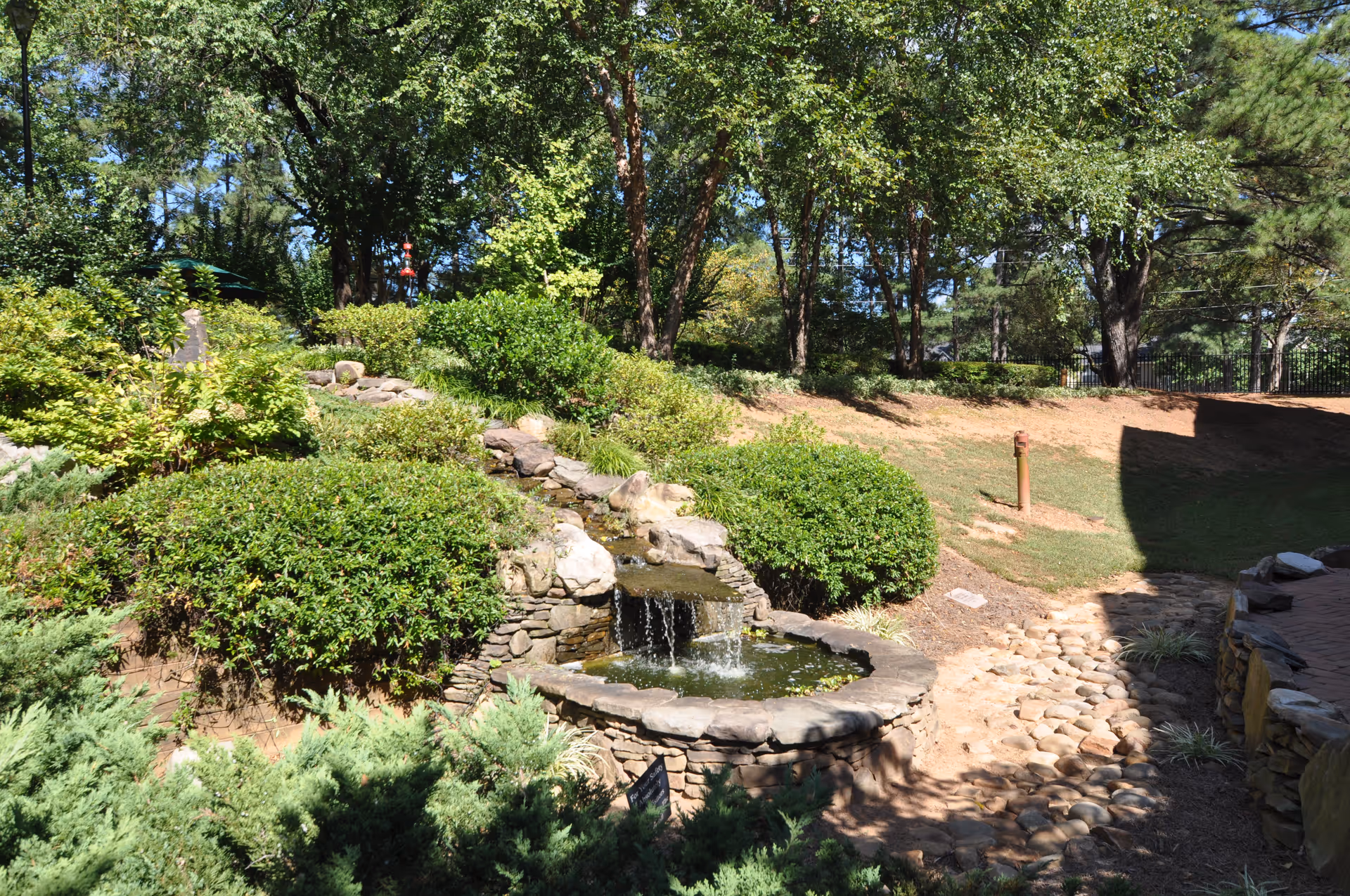 A landscaped outdoor garden area with a small stone waterfall flowing into a pond surrounded by green bushes and trees under a clear blue sky.