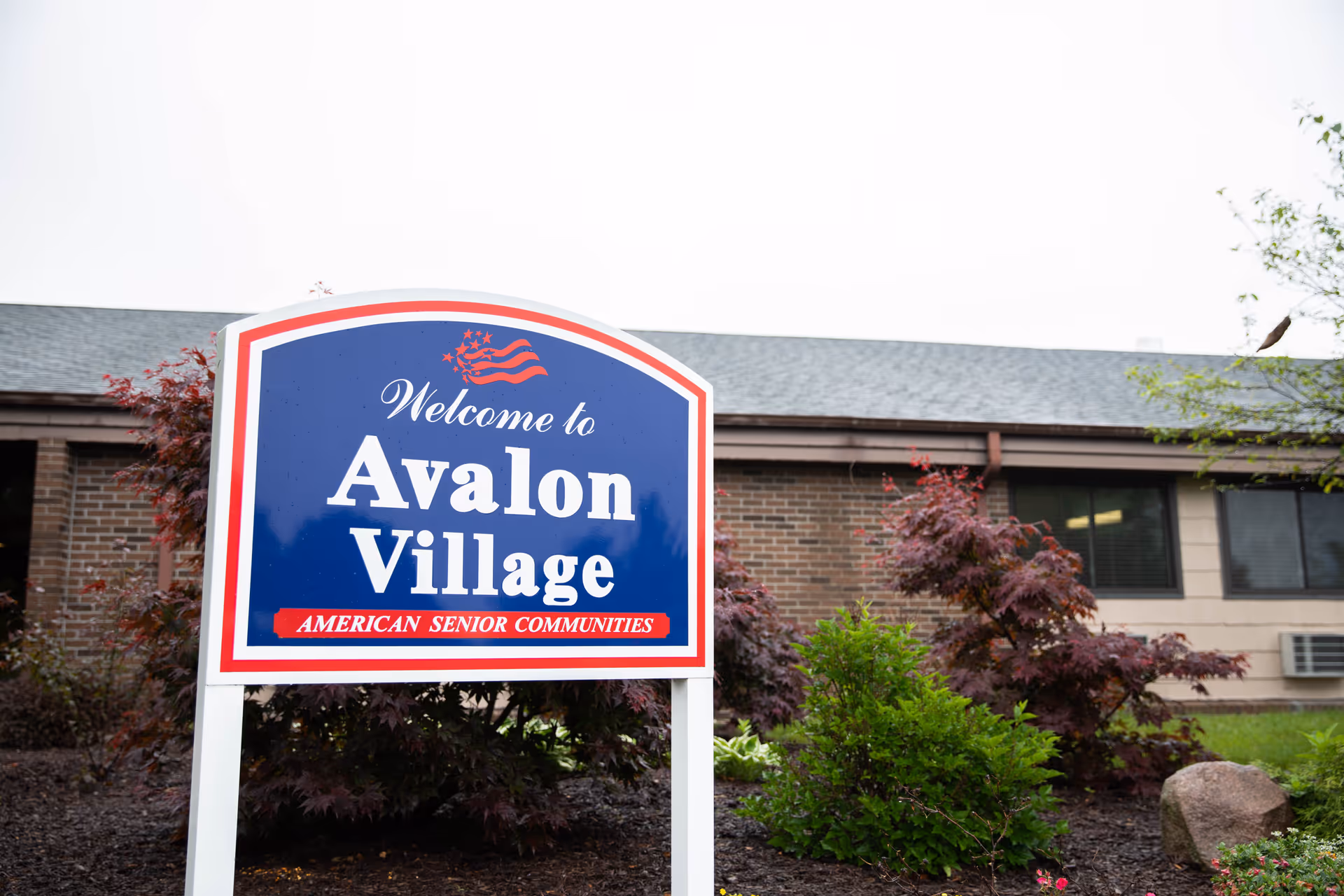 A blue and white sign with red accents that reads 'Welcome to Avalon Village American Senior Communities' in front of a brick building with windows and surrounded by bushes and small trees.