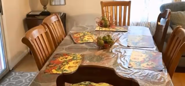 A dining table with a clear plastic cover and floral placemats, surrounded by wooden chairs. A decorative fruit basket is placed in the center of the table. The room has a beige wall, a side table with a lamp, and a window with sheer curtains allowing natural light.