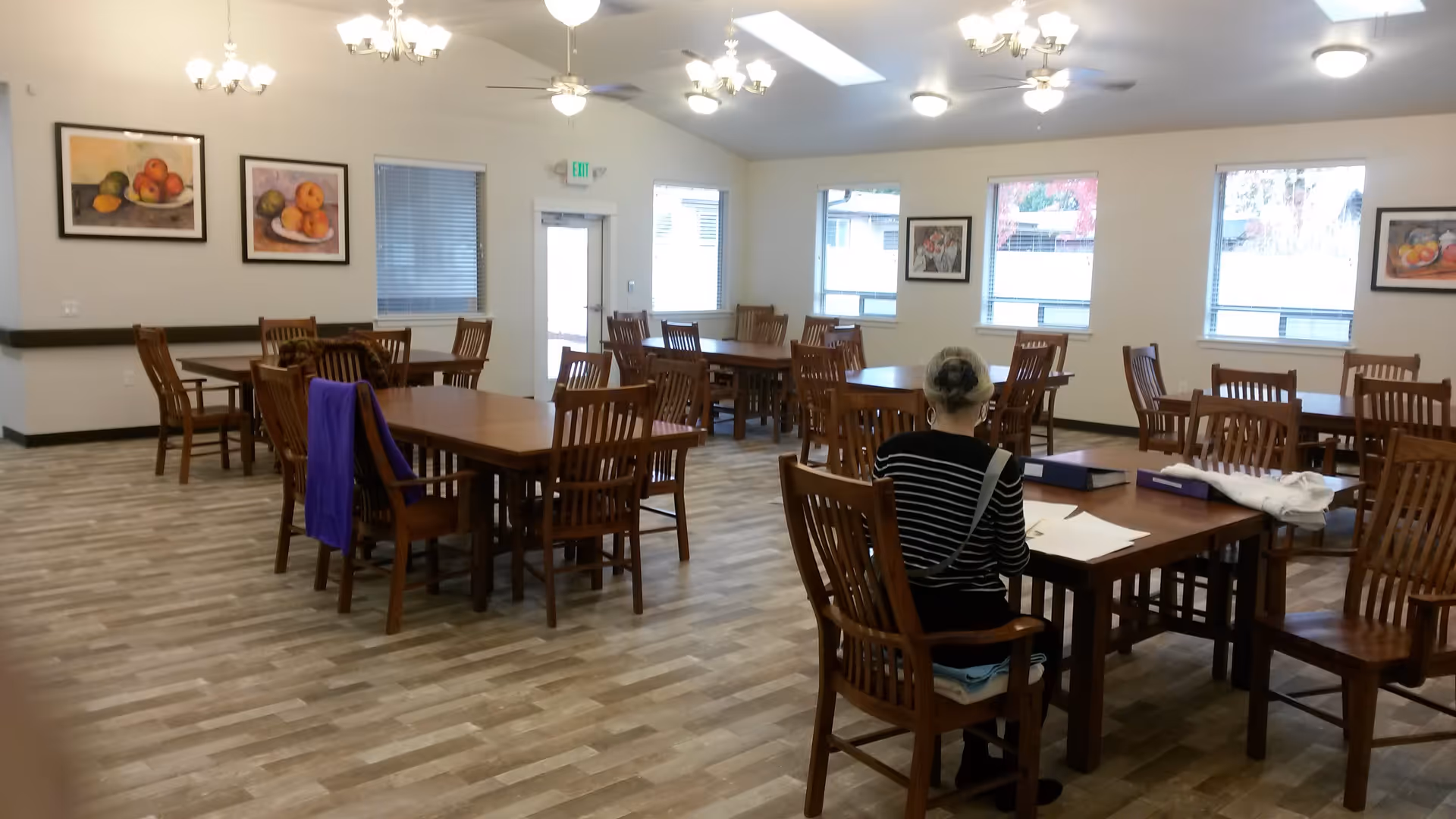 A spacious dining room with multiple wooden tables and chairs arranged neatly. A person with gray hair tied back is sitting at one of the tables, facing away, with papers and binders on the table. The room has several windows letting in natural light, framed fruit paintings on the walls, and ceiling lights with fans.