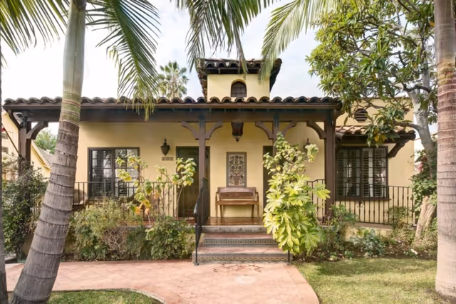 Mediterranean-style single-story house front with a covered porch, tiled roof, palm trees, and landscaped plants.