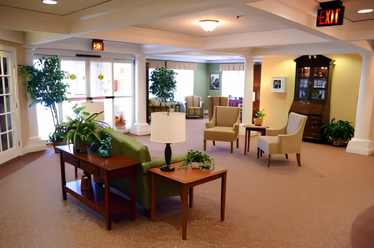 A spacious and well-lit senior living facility common area with beige carpeting, several armchairs, a green sofa, wooden tables with lamps and plants, large windows, and potted plants. An exit sign is visible on the ceiling.