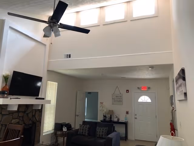 High-ceiling communal living room with a ceiling fan, TV above a stone fireplace, seating and the front entrance.