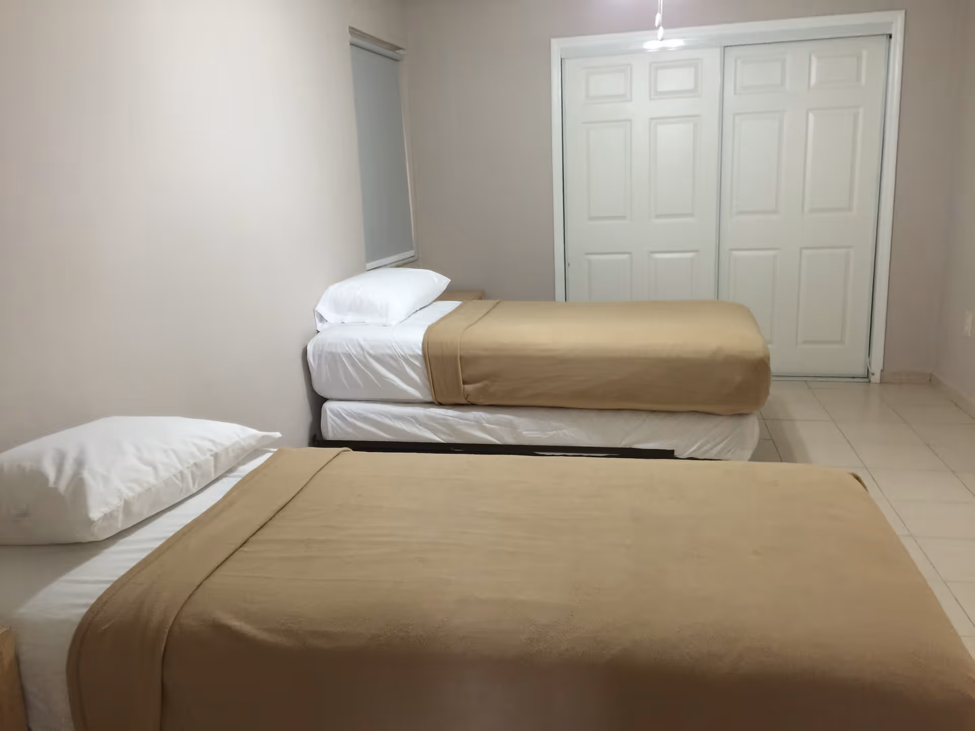 A simple bedroom with two single beds placed parallel to each other, each made with white sheets, white pillows, and beige blankets. The room has light-colored walls, a window with a closed blind, and a white double-door closet at the far end. The floor is tiled in a light color.