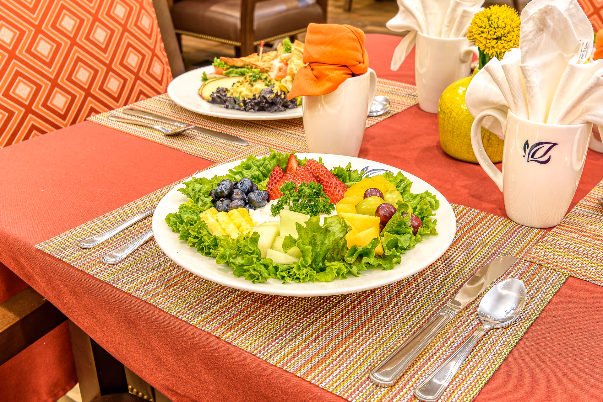 Plate of mixed fruit and salad on a set table with utensils, napkins, and mugs in a dining room.