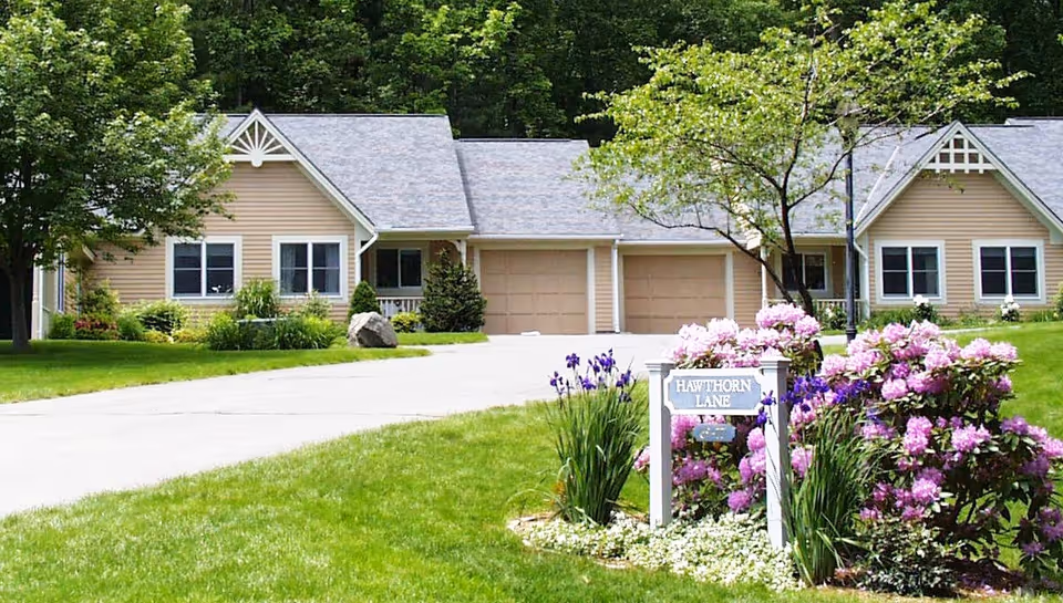 Front view of single-story beige retirement cottages with garages, driveway, green lawn and a flowerbed sign reading 'Hawthorn Lane'.