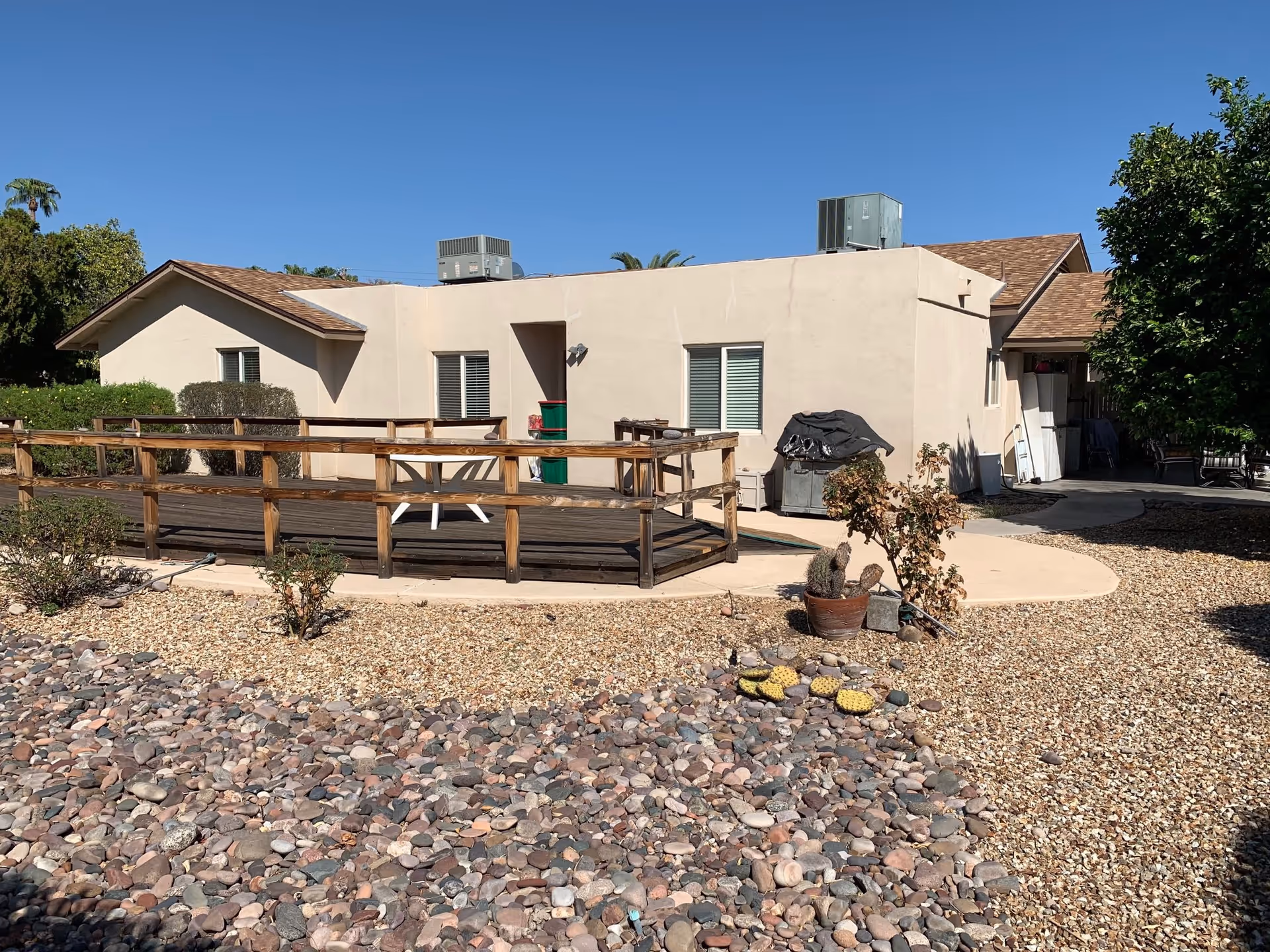 Exterior view of a single-story building with beige walls and a brown shingled roof under a clear blue sky. The building has a wooden ramp with railings leading to a door. The surrounding yard is covered with rocks and gravel, with a few small plants and potted cacti. There are two air conditioning units on the roof and some outdoor furniture and equipment near the building.