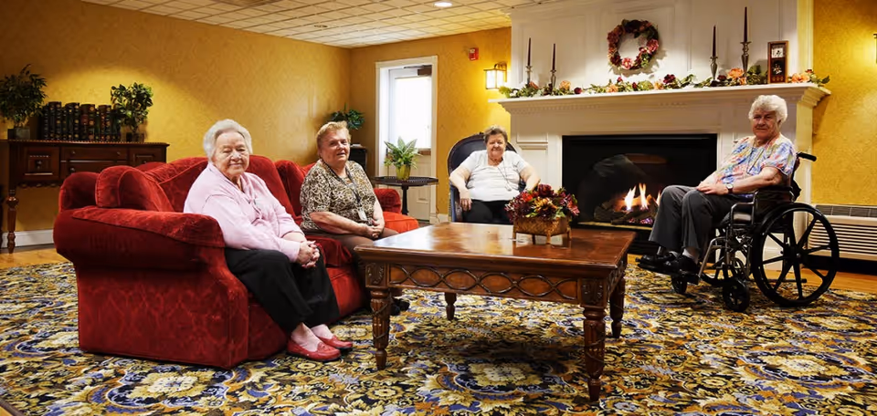 Four older adults seated in a carpeted common room with a red sofa, wooden coffee table and a lit fireplace.