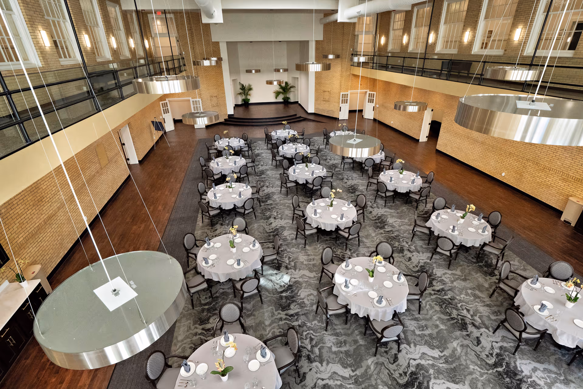 A large elegant dining/event hall with multiple round tables set for service, viewed from an upper balcony.