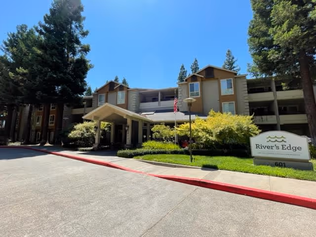 Exterior view of River's Edge senior living facility showing a multi-story building with beige and brown walls, surrounded by tall pine trees and landscaped bushes. The entrance has a covered driveway, and an American flag is visible near the entrance. A sign with the facility name 'River's Edge' and the number 601 is in the foreground on the right side.