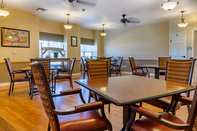 A well-lit dining room with multiple wooden tables and cushioned chairs arranged neatly. The room has light-colored walls, three windows with blinds, ceiling fans, and hanging light fixtures. There is a framed picture on one wall and a door on the right side.