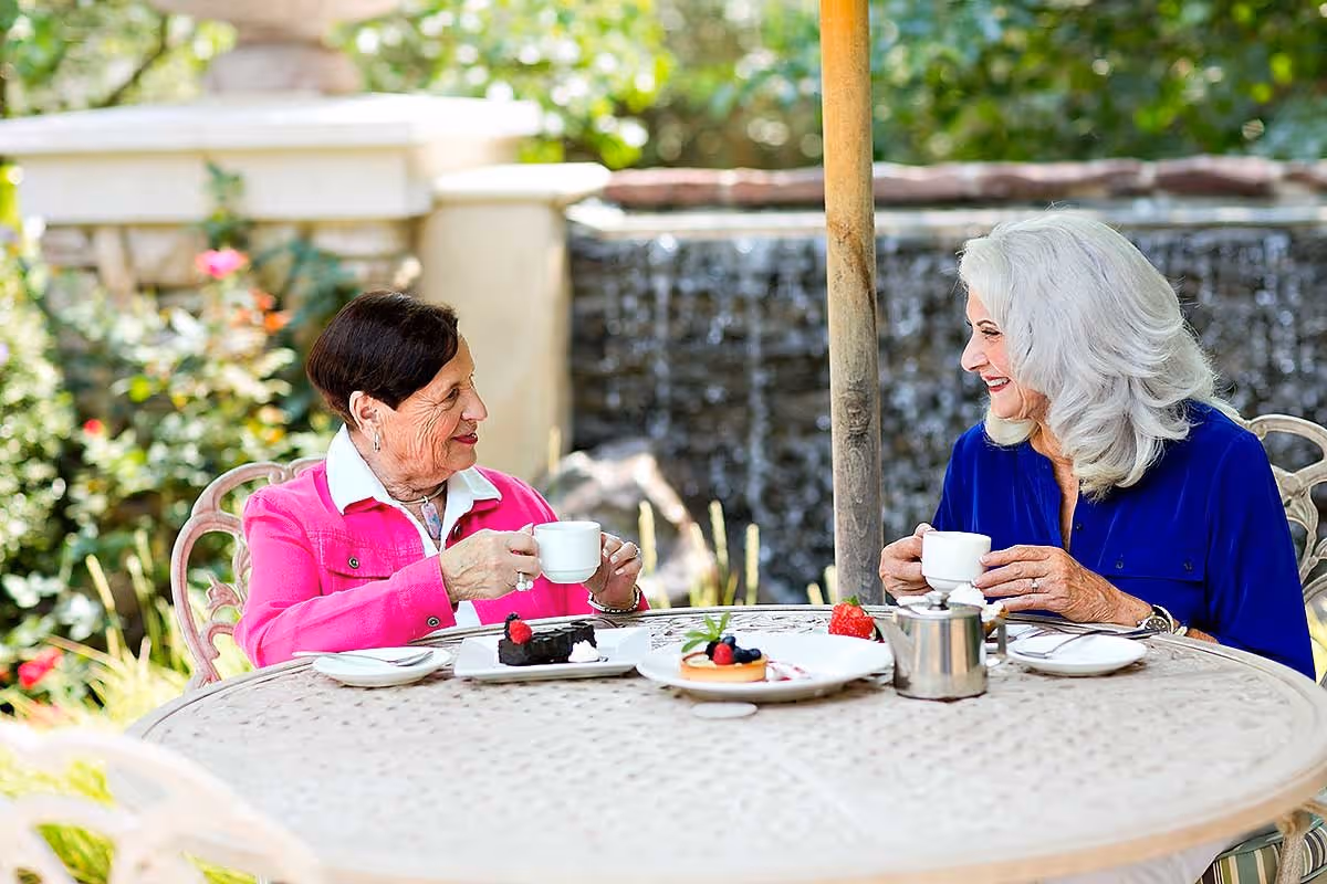 Two older women sit at a round outdoor table drinking tea and enjoying desserts in a garden setting.