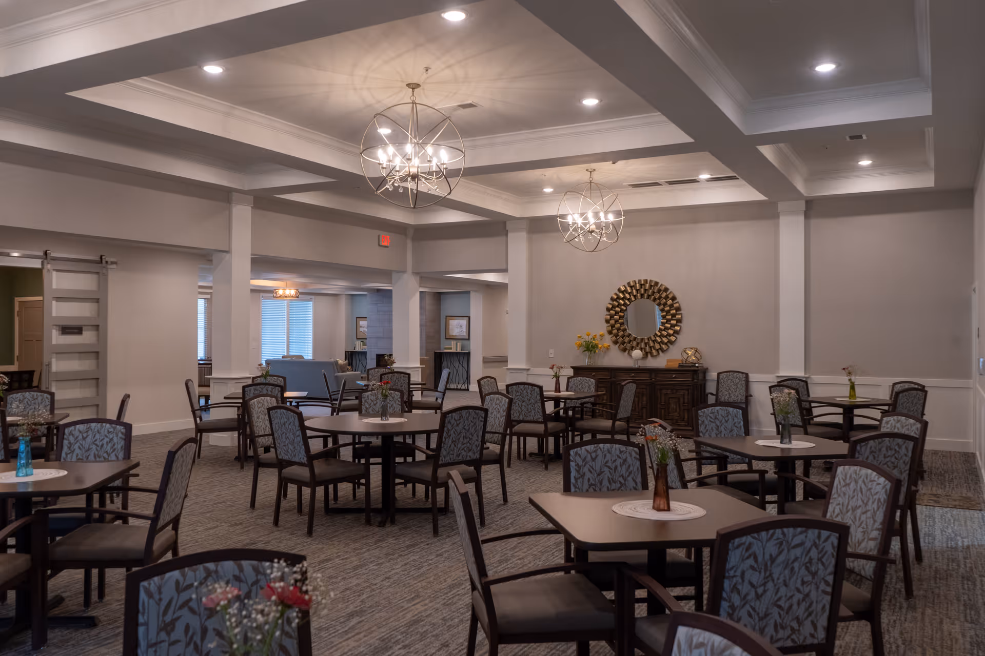 A spacious dining room with multiple round tables and chairs arranged neatly. Each table has a small vase with flowers. The room features modern chandeliers hanging from a coffered ceiling, neutral-colored walls, and a decorative mirror above a wooden sideboard against the far wall.