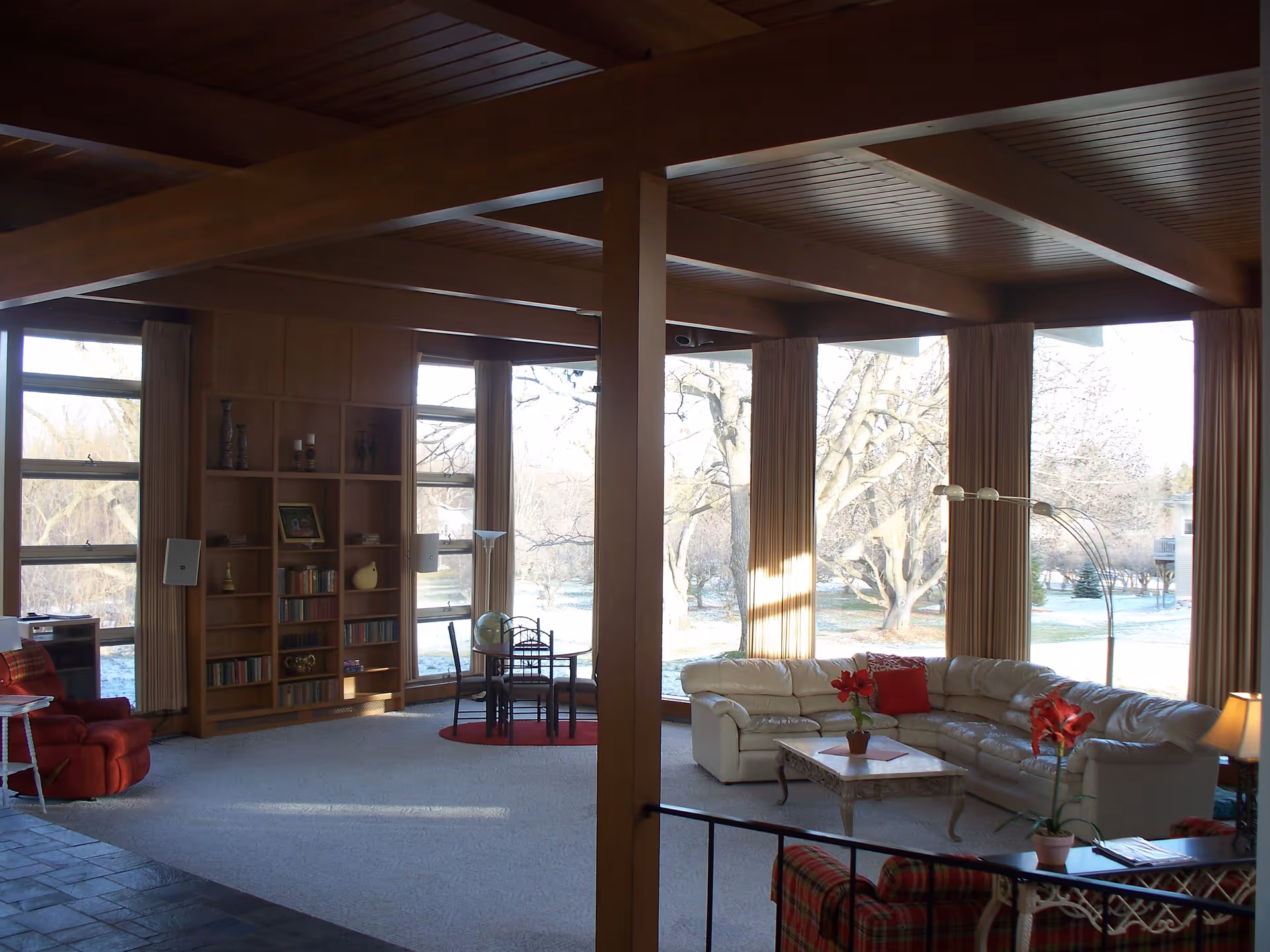 Bright living room with a wood-beamed ceiling, floor-to-ceiling windows, a cream sectional sofa and built-in bookshelves.