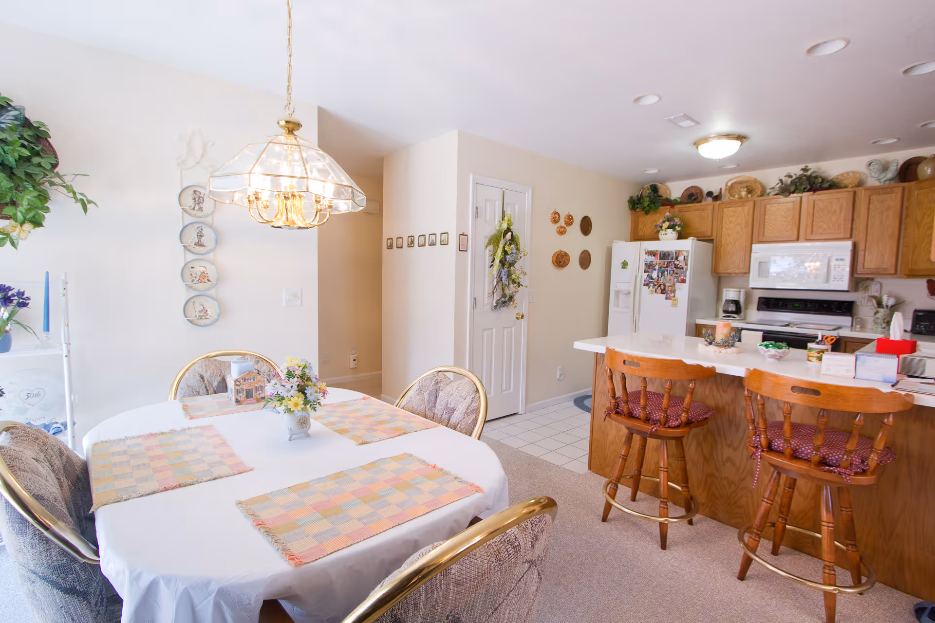 Bright dining area with a table set with placemats and a hanging chandelier next to an open kitchen with a breakfast bar and wooden stools.