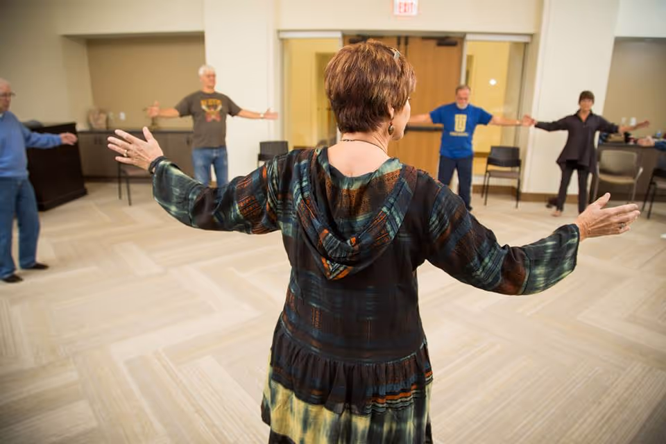 A group of elderly people standing in a circle in a spacious room, participating in a stretching or exercise activity led by a woman with short hair wearing a patterned hoodie, with arms extended outward.