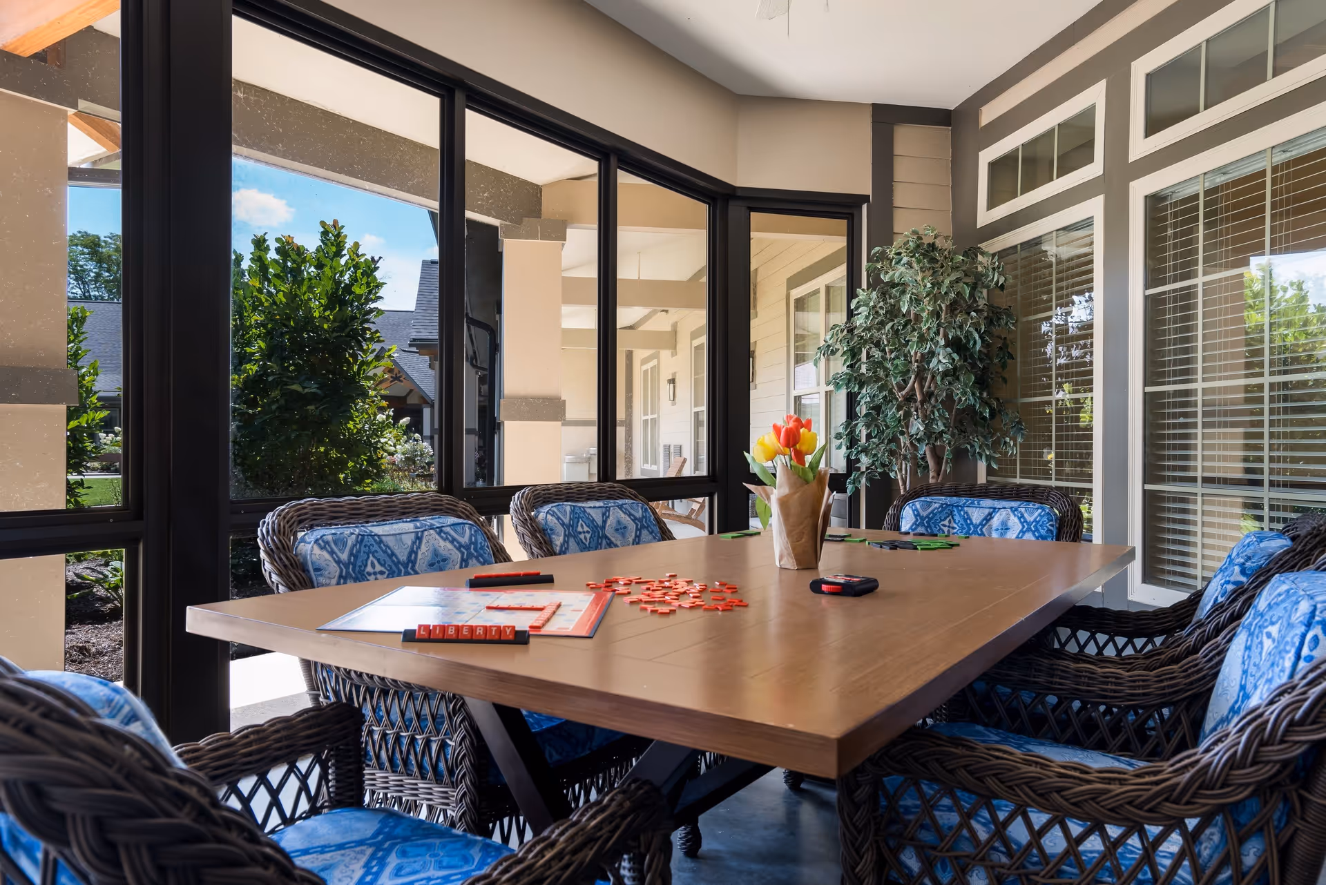 A cozy indoor seating area with a wooden table surrounded by wicker chairs with blue patterned cushions. On the table, there is a Scrabble game in progress with tiles spelling 'LIBERTY' and a vase with colorful tulips. Large windows provide a view of the outdoor garden area with greenery and a covered walkway.