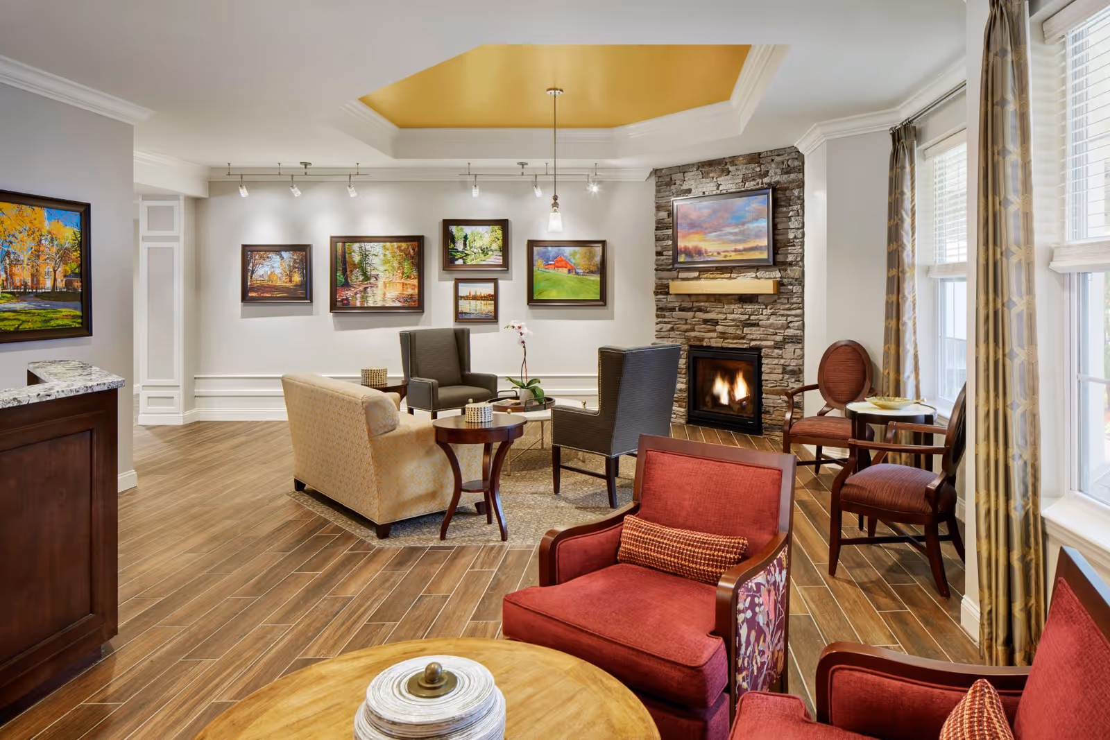 Cozy senior living common room with upholstered chairs and sofas arranged around a coffee table facing a stone fireplace and wall art.